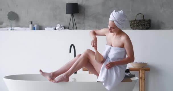 Halv naken vacker kvinna insvept i badhandduk med hjälp av en pensel massager, gör utjämning anti-celluliter kroppsmassage på benen sitter på kanten av badkar samtidigt förbereda avkopplande spa bubbelbad — Stockvideo
