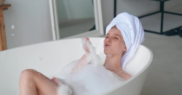 Красива жінка з рушником для ванни на голові лежить на ванні, посміхається, розслабляється, масажує її тіло, приймаючи бульбашкову ванну з піною у ванній кімнаті білого дому, насолоджуючись вихідним днем. Концепція догляду за тілом — стокове відео