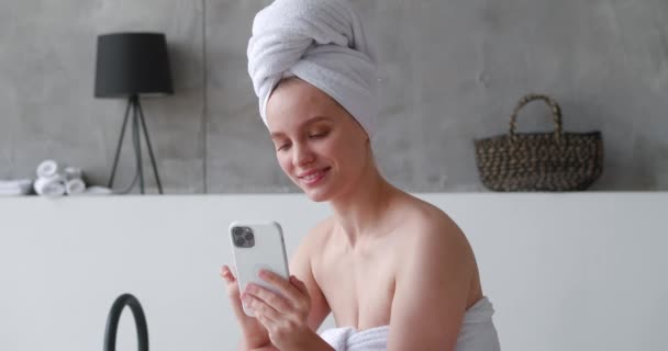 Close-up retrato de mulher bonita semi-nua envolto em toalha de banho, usando um telefone celular, mensagens de texto, deslizando enquanto sentado na banheira do banheiro doméstico moderno, antes de tomar banho relaxante com espuma — Vídeo de Stock