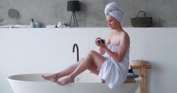 Halfnaakte mooie vrouw, gewikkeld in witte badhanddoeken, brengt vochtinbrengende crème aan op haar benen, maakt een gladde massage, zittend aan de rand van het bad tegen de achtergrond van het water dat uit de kraan stroomt — Stockvideo