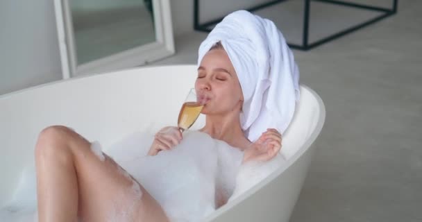 Attractive blonde woman with closed eyes of pleasure, wrapping head in white terry bath towel, lying in bathtub and enjoying bath with foam, tasting delicious white sparkling wine at home at weekends — Stock Video