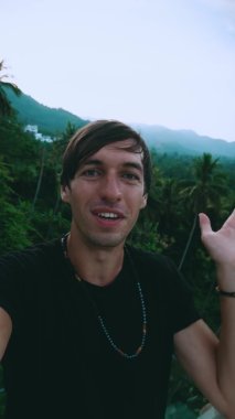 Yeşil orman arka planında selfie ya da video görüşmesi yapan gülümseyen genç turistin dikey video portresi. Genç erkek, görüntülü sohbet için tropikal bir ülkeyi geziyor..