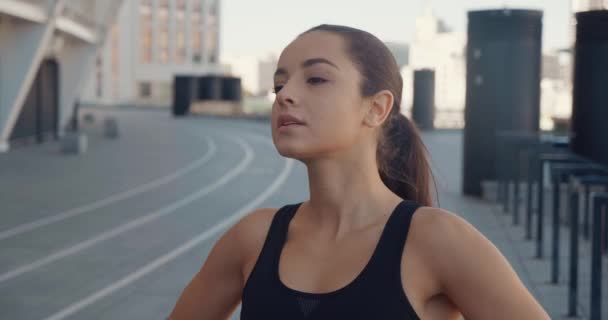 Portret van jonge mooie atletische vrouw in sportkleding buiten staan op stedelijke achtergrond, glimlachen en poseren voor de camera, terwijl rusten na de ochtend run — Stockvideo