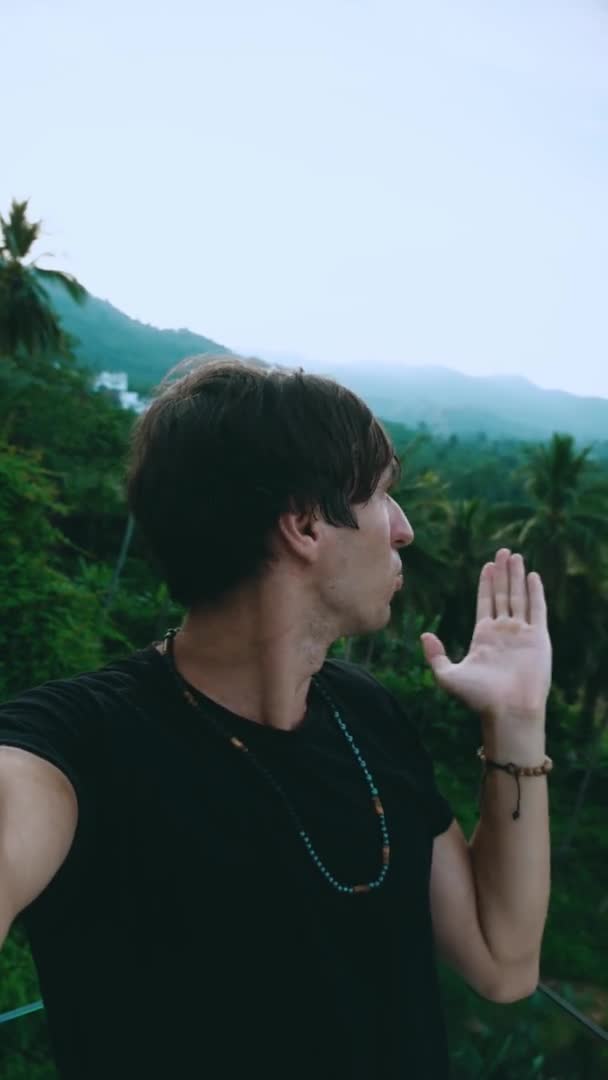 Vertikales Videoporträt eines lächelnden jungen Mannes, der ein Selfie oder einen Videoanruf auf grünem Dschungel-Hintergrund macht. Junger Mann reist durch tropisches Land und macht Videochat. — Stockvideo
