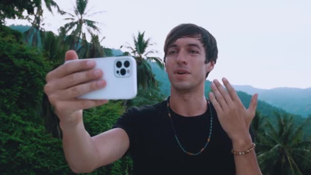 Hombre feliz viajero haciendo videollamadas en tiempo real con teléfono inteligente al aire libre en el hermoso fondo de la selva, saludando a la gente en la pantalla del teléfono. Joven blogger haciendo videoblog vlog usando teléfono móvil — Vídeo de stock