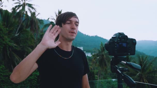 Улыбающийся блогер разговаривает с камерой, записывает видео блог с помощью профессиональной видеокамеры, установленной на штативе на фоне гор в джунглях — стоковое видео