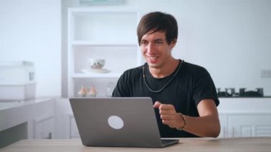 Mutfakta dizüstü bilgisayarı olan heyecanlı genç bir adam. Mutlu serbest çalışan erkek jest yapıyor. Piyangoyu kazandıktan sonra heyecanlı bir şekilde dizüstü bilgisayara bakıyor. Evde mutfaktaki masada oturuyor.