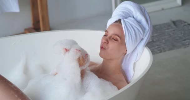 Красива спокійна, в захваті, задоволена приваблива жінка з білим махровим рушником на голові, насолоджується бульбашковою ванною вдома, грає з мильною піною, дме бульбашки, розслабляється у вихідний день вдома спа — стокове відео