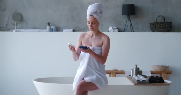 Houkutteleva nainen kääritty valkoinen kylpy pyyhe istuu reunalla kylpyamme ristissä paljain jaloin, avasi pullon, jossa on kauneustuote ja soveltamalla kosteusvoidetta hänen ruumiinsa tekee tasoitus hieronta — kuvapankkivideo