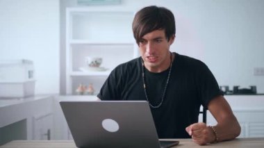 Öfkeli sinirli adam bağırıyor ve dizüstü bilgisayarında gördüğü şeye agresif bir tepki veriyor. Öfkeyle yumruğunu masaya vuruyor. Evde mutfak masasında otururken.