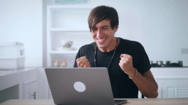 Dizüstü bilgisayara bakan heyecanlı serbest çalışan adam online bahsi kazandı. Şanslı genç erkek harika haberler okuyor ve evdeki mutfakta bilgisayar başında çalışırken evet hareketi yapıyor.