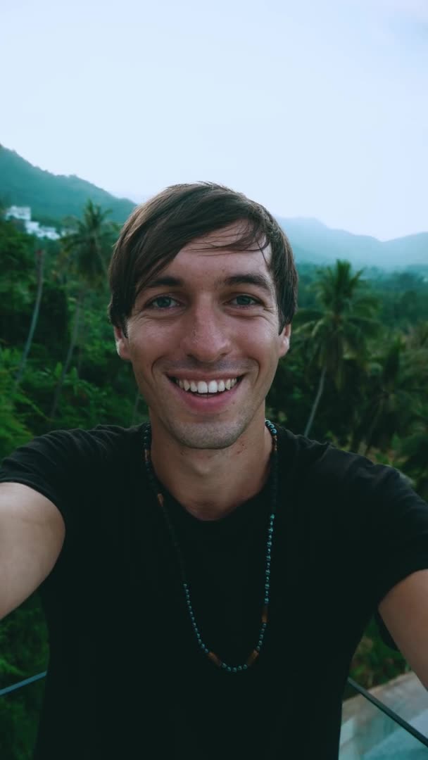 Вертикальний відео-портрет усміхненого молодого чоловіка, який робить селфі або відеодзвінок на фоні зелених джунглів лісу. Молодий чоловік подорожує по тропічній країні робить відеочат . — стокове відео
