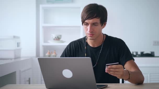 Junger Mann beim Online-Einkauf mit Kreditkarte und Laptop in der Küche zu Hause. Lächelnder Mann genießt Internet-Einkauf von zu Hause aus mit Kreditkarte — Stockvideo