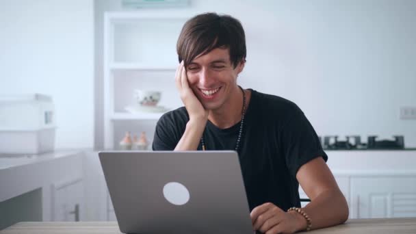 Молодой счастливый человек смотрит на монитор ноутбука и смеется, читая смешные новости, сидя дома за кухонным столом. Улыбающийся мужчина смотрит смешные видео в Интернете и смеется над тем, что он видел — стоковое видео