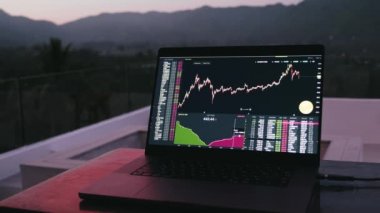 Masanın üzerinde duran dizüstü bilgisayar, borsa, çevrimiçi ticaret, kripto para birimi görüntüsü orman dağlarında günbatımında arka planda..