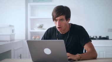 Şok olmuş erkek, dizüstü bilgisayardan kötü haberler izliyor ve evdeki mutfak masasında otururken ağzını dehşet içinde kapatıyor. İş adamı ofisteki bilgisayardan kötü sonuçlar alıyor..