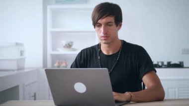 Serbest çalışan genç iş adamı dizüstü bilgisayarı açıyor ve evdeki mutfak masasında otururken bilgisayarın başında çalışmaya başlıyor. Ev işyerinde. İçişleri Bakanlığı.