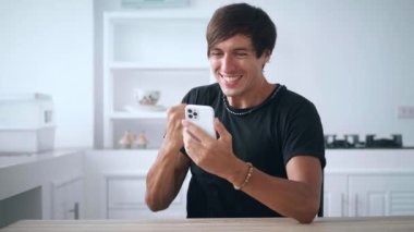 Akıllı telefona bakan heyecanlı adam online bahis kazanma başarısını kutluyor. Şanslı genç erkek cep telefonundan iyi haberler okuyor ve mutfak masasında evet işareti yapıyor.