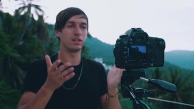 Genç blogcu kameraya konuşuyor, profesyonel bir video kamera kullanarak video blogu kaydediyor. Ormandaki dağların arka planındaki bir tripoda monte edilmiş.