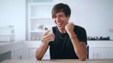 Akıllı telefona bakan heyecanlı adam online bahis kazanma başarısını kutluyor. Şanslı genç erkek cep telefonundan iyi haberler okuyor ve mutfak masasında evet işareti yapıyor.