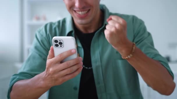 Close-up vzrušený muž při pohledu na smartphone oslavit úspěch mobilní vyhrát on-line sázku. Šťastný mladý muž čte dobré zprávy na mobilním telefonu a dělat ano gesto u stolu domácí kuchyně — Stock video