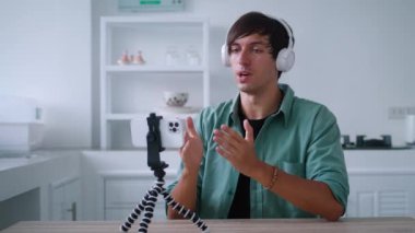 Kablosuz kulaklıklı genç adam cep telefonuyla bir video görüşmesinde konuşuyor. Popüler blogcu evinde, akıllı telefonunu kullanarak internet için bir video blogu kaydediyor.