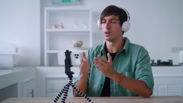 Young man in wireless headphones is talking on a video call using his mobile phone. Popular blogger is recording a video blog for the internet using his smartphone, at home in the kitchen — Vídeos de Stock
