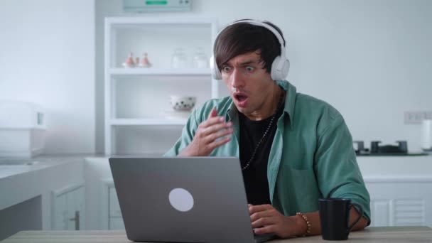 Scared young man in headphones getting bad results on computer at home office. Young male watching bad news on a laptop and covering his mouth in horror while sitting at the kitchen table at home — Vídeos de Stock