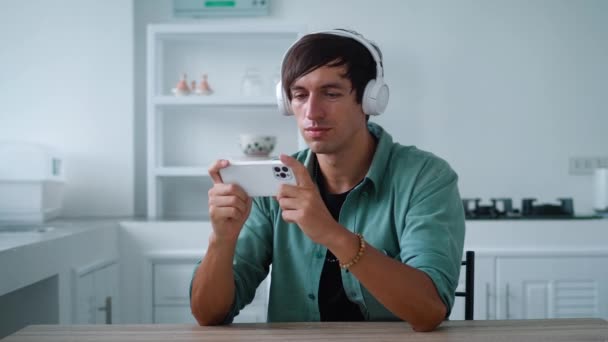 Молодой человек в наушниках играет в игру на своем смартфоне, сидя дома на кухонном столе — стоковое видео