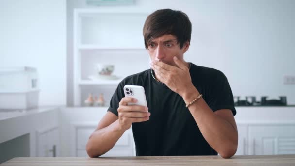 Shocked young man using smartphone, loses, dissatisfied with bad news at home. Upset depressed male holds mobile phone in hands on background kitchen. Mobile losing concept — Vídeo de Stock