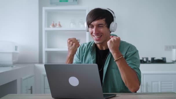 Excited man freelancer in headphones looking at laptop celebrate success win online bet. Lucky young male reading great news and making yes gesture while working at computer at home kitchen — 비디오