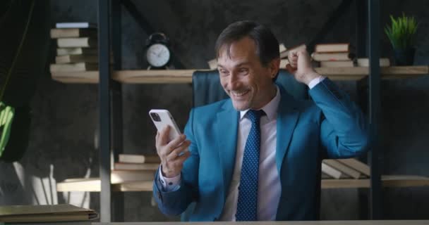 Amazed mature business man receiving message offer opportunity, happy boss reading good news in smartphone, sitting at office. Excited overjoyed male celebrating success mobile phone victory — Stock Video