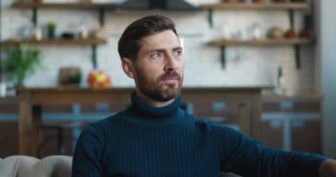 Kanepede otururken kameraya bakarak gülümseyen başarılı beyaz adamın yakın plan portresi. Mutfak arka planında sakallı bir erkek resmi var.