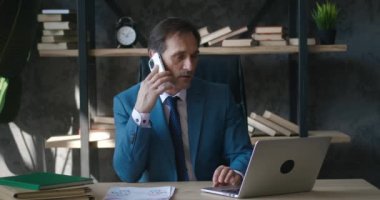 Serious middle-aged business man conducts business negotiations using a mobile phone while working at a computer in a modern office. Classic suit dressed