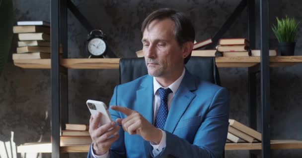 Professional senior business man holding modern smartphone texting message in office. Boss using modern mobile phone technology business app, typing email sitting at work desk. — Wideo stockowe