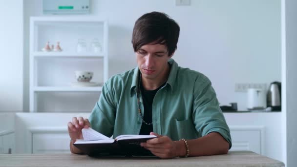 Młody mężczyzna siedzi przy kuchennym stole w domu i czyta książkę, przeszukując strony. — Wideo stockowe