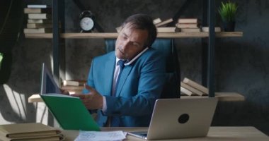 Çoklu görev yapan orta yaşlı iş adamı, iş belgeleriyle çalışırken ve modern ofiste dizüstü bilgisayar kullanırken cep telefonu kullanarak iş görüşmelerinde bulunuyor. Pek çok görevi yerine getirir. Klasik takım elbise.
