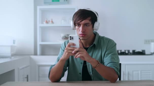 Happy man in headphones looking at smartphone celebrate success mobile win online bet. Lucky young male reading good news on mobile phone and making yes gesture at home kitchen table — Stock video