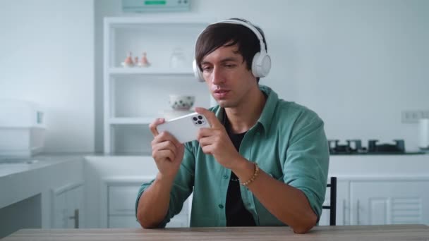 Hombre joven en auriculares jugando juego de aplicación en su teléfono inteligente mientras está sentado en la mesa de la cocina en casa — Vídeo de stock