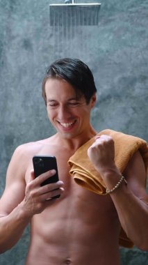 Akıllı telefona bakıp banyo yapmadan önce banyonun yanında duran havluyla sevinen heyecanlı bir adamın dikey video portresi. Jest evet. Mobil kazanç kavramı