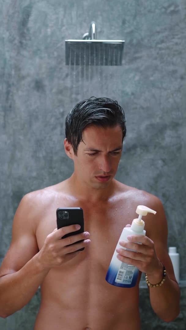 Vertical video of a man looking for information on the Internet about body care using a smartphone, standing in the bathroom on the background of the shower. Self care concept — Stockvideo