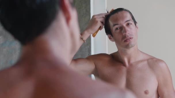 Young man combs his hair and does his morning routine, self-care in the bathroom, looking at himself in the mirror. — Vídeo de Stock