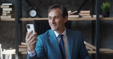 Olgun iş adamı akıllı telefondan video görüşmesi yapıyor. Resmi takım elbiseli kıdemli erkek CEO modern ofiste otururken cep telefonuyla selfie görüntülü sohbet yapıyor..