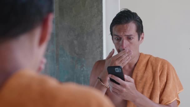 Worried man looking at smartphone and shocked by bad news, covering his mouth with his hand in horror, standing in the bathroom, feeling stressed, being in the bathroom on against the mirror — Vídeos de Stock