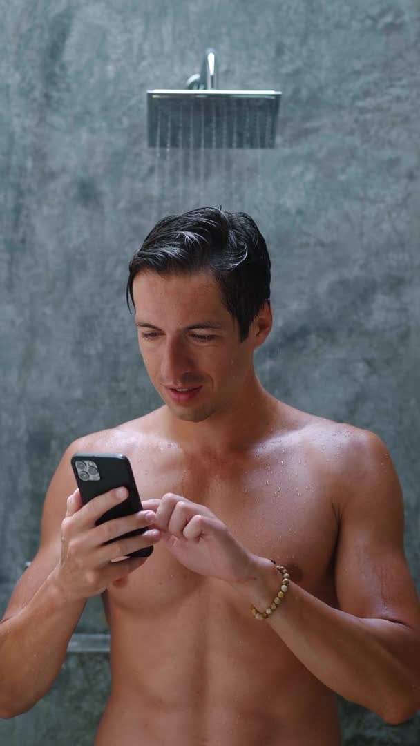 Video verticale ritratto di giovane uomo sorridente si trova nel bagno vicino alla doccia e utilizzando smartphone app prima del bagno. Concetto di auto cura — Video Stock