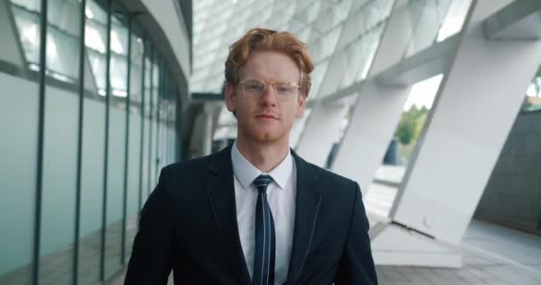 Portrait of ambitious red-haired man top manager looking at the camera and smiling. Young male businessman in formal suitn and eyeglasses standing in the business district near the office building — Vídeos de Stock