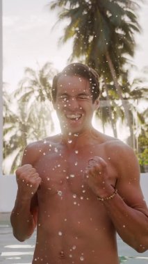 Vertical video portrait of smiling excited young man looking at camera and rejoicing in a win or good news, making yes gesture, standing outdoors in resort hotel by the pool at a beautiful sunset