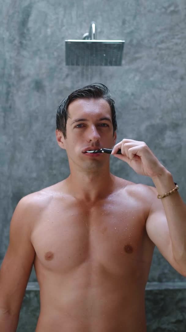 シャワーの背景にあるバスルームに立っている間に彼の歯を磨く若いハンサムな男の垂直ビデオ、朝のルーチン — ストック動画