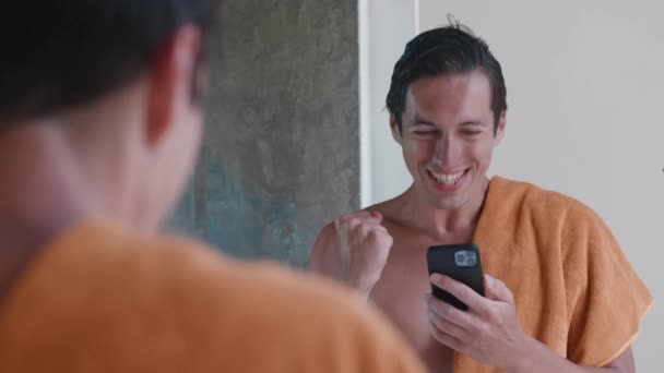 Podekscytowany uśmiechnięty mężczyzna patrzący na smartfona i cieszący się z dobrych wieści z ręcznikiem stojącym w łazience przed lustrem przed kąpielą. Tak, zgadza się. Mobilna koncepcja wygranej — Wideo stockowe