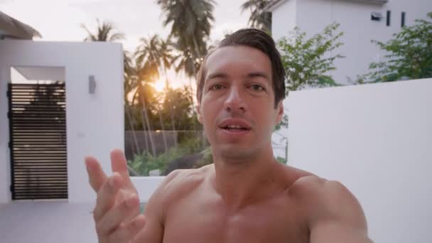 POV young man holding mobile phone and making selfie video calling while holidays. Popular blogger talking to camera, recording a video blog for the internet outdoors at luxury pool villa. — Wideo stockowe